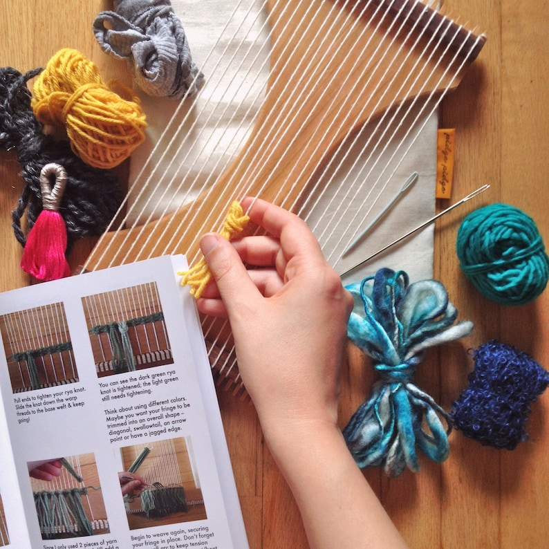 May include: A craft image featuring a wooden weaving loom with white warp threads. A hand holds yellow yarn, with various colored yarn balls and a needle nearby. A book with weaving instructions is open.