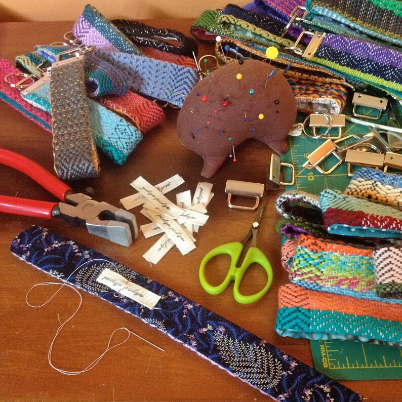 May include: A collection of handmade woven fabric straps in various colors and patterns, along with sewing tools. The straps are in shades of blue, green, orange, and purple. Tools include scissors, pliers, a pincushion, and metal hardware.
