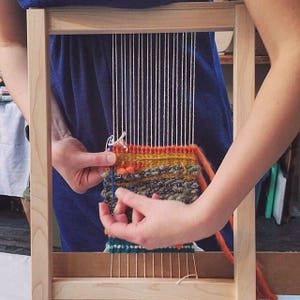 May include: A wooden weaving loom with a partially completed textile. The textile features a mix of orange, yellow, green, and blue yarns. The weaver's hands are visible, working with the threads. The loom is rectangular and made of light-colored wood.