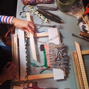 May include: A top-down view of a craft project in progress, featuring wooden weaving looms. The looms hold woven yarn in shades of gray, red, and green. Various craft supplies, including yarn, scissors, and a fork, are scattered around the looms.