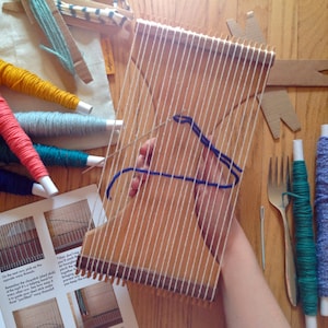 May include: A wooden weaving loom with white warp threads is held by a person. Blue yarn is being woven through the loom. Various colored yarn spools, a metal fork, and a needle are on the wooden surface, along with instructions.