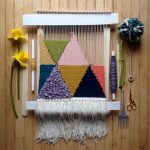 May include: A colorful geometric woven wall hanging in progress, featuring triangles in shades of pink, green, gold, teal, and navy. The weaving is on a wooden loom, with white fringe at the bottom. Additional craft supplies include yarn, scissors, and a comb.
