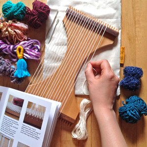 May include: A wooden weaving loom with white threads, surrounded by colorful yarn in teal, purple, and burgundy. A hand is holding a white thread, and a booklet with instructions is visible.