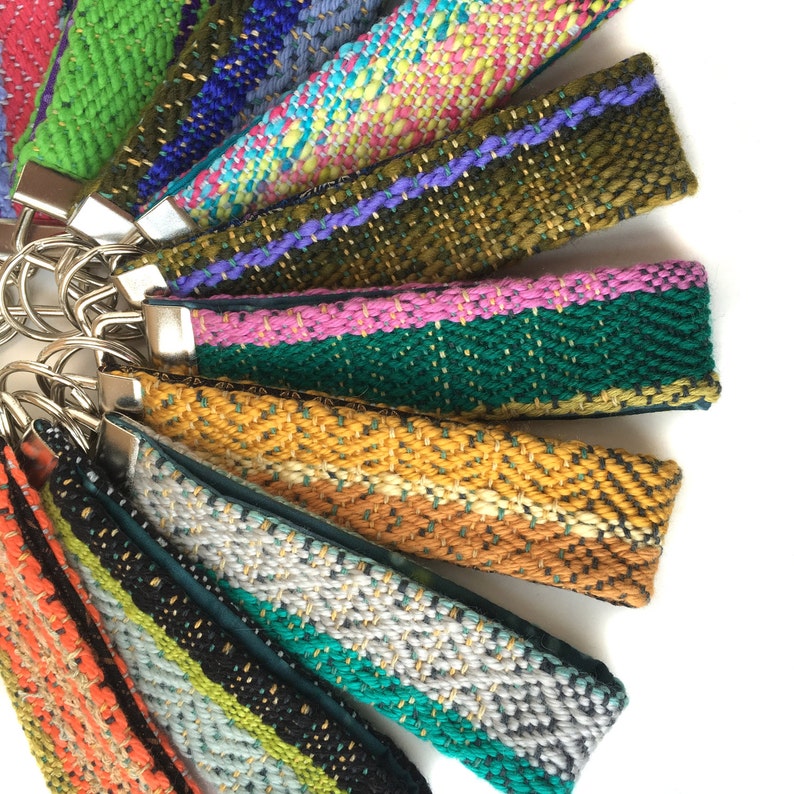 May include: A collection of colorful woven wrist keychains with silver-tone hardware. The keychains feature various patterns and colors, including green, blue, pink, and yellow. Each keychain is approximately 6 inches long.