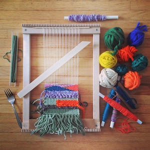 May include: A wooden weaving loom with a partially woven textile in shades of green, purple, orange, and blue. Various balls of yarn in green, yellow, orange, and blue are scattered around the loom, along with weaving tools.