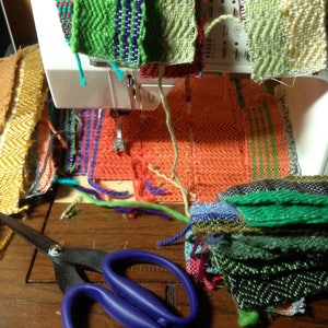 May include: A sewing machine with colorful woven fabric samples in various patterns and colors. A pair of scissors with blue handles rests on a wooden surface near the fabric. The fabrics include green, orange, and red hues.