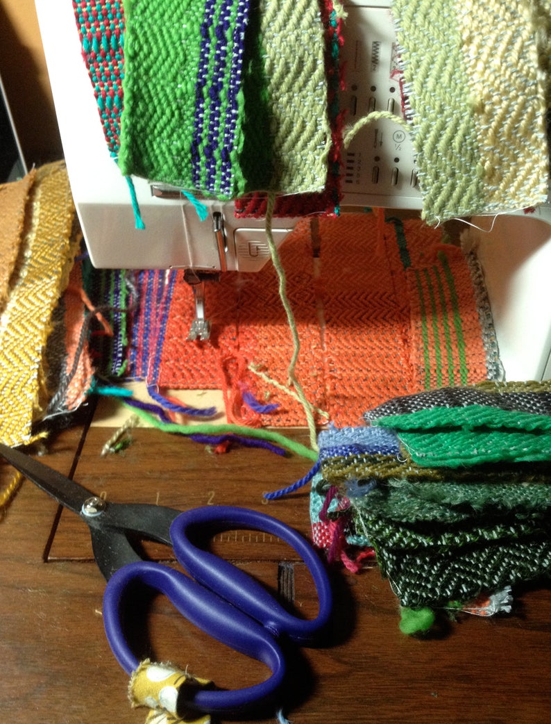 May include: A sewing machine with colorful woven fabric samples in various patterns and colors. A pair of scissors with blue handles rests on a wooden surface near the fabric. The fabrics include green, orange, and red hues.