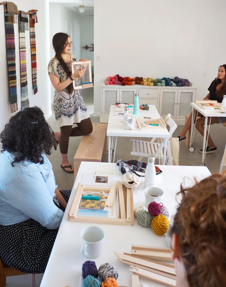 May include: A craft class in a bright room. A woman holds a small wooden weaving loom, demonstrating to a group. Colorful yarn balls and weaving projects are on the tables. Woven wall hangings in various colors are displayed on the wall.