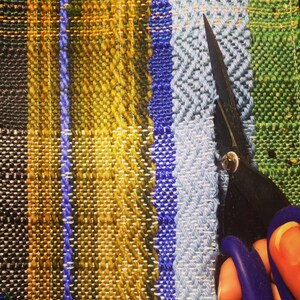 May include: Close-up of a colorful woven textile with vertical stripes in shades of black, yellow, blue, and green. A pair of black scissors with blue handles is cutting the fabric.