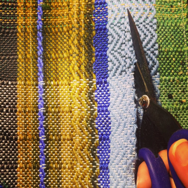May include: Close-up of a colorful woven textile with vertical stripes in shades of black, yellow, blue, and green. A pair of black scissors with blue handles is cutting the fabric.