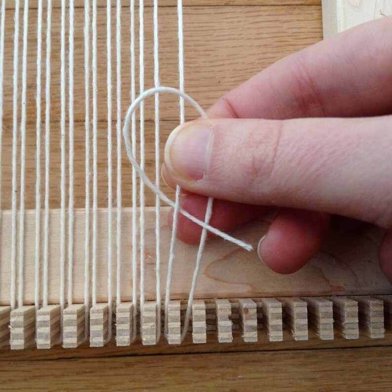 May include: Close-up of a wooden weaving loom with white yarn. A hand is shown manipulating the yarn, creating a loop. The loom has vertical threads, ready for weaving. The wood has a natural, light brown color.