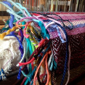 May include: Close-up of a colorful woven textile in progress on a loom. The piece features a mix of vibrant yarns in shades of blue, pink, red, and green. The fringe of the textile is visible, showcasing the individual strands of yarn.
