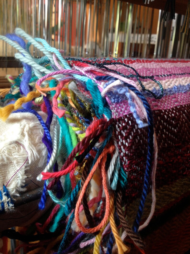 May include: Close-up of a colorful woven textile in progress on a loom. The piece features a mix of vibrant yarns in shades of blue, pink, red, and green. The fringe of the textile is visible, showcasing the individual strands of yarn.