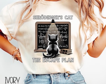 May include: Ivory-colored t-shirt featuring a black and white illustration of a cat in front of a chalkboard with equations. The text "SHRÖDINGER'S CAT THE ESCAPE PLAN" is printed on the shirt. The shirt is made of a soft material.