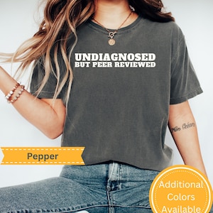 May include: A gray t-shirt with the text "Undiagnosed but Peer Reviewed" in white. The shirt is being worn by a person with long brown hair. The shirt is cropped and the person is wearing blue jeans.