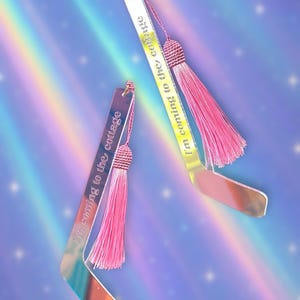 May include: Two mirrored acrylic bookmarks with pink tassels. Each bookmark has the phrase "I'm coming to the cottage" etched on it. The bookmarks are in a hockey stick shape and reflect a rainbow of colours.