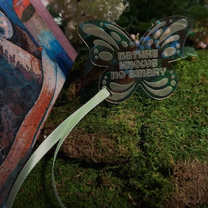 Può includere: Un ornamento in acrilico traslucido a forma di farfalla con il testo "nature knows no binary" in un carattere stilizzato. L'ornamento è fissato a un nastro verde chiaro e presenta un sottile riflesso iridescente sulla superficie.