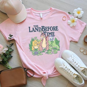May include: A light pink t-shirt with a graphic from "The Land Before Time" and the text "The Land Before Time". The shirt is styled with white trainers, a pink baseball cap, and a brown purse. The shirt is knotted at the waist.