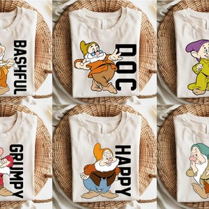 May include: Six cream-coloured t-shirts featuring cartoon images of Snow White's dwarfs. Each shirt displays a different dwarf with their name in bold black letters: Bashful, Doc, Dopey, Grumpy, Happy, and Sleepy. The shirts are displayed on woven placemats.