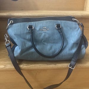May include: A Coach handbag with a denim-like blue fabric body and black leather trim. The bag has a top zipper closure, dual handles, and a detachable shoulder strap. The Coach logo is visible on the front.