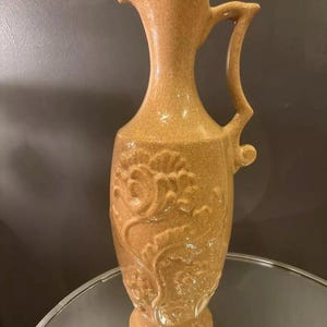 Puede incluir: Una jarra de cerámica marrón alta con una superficie texturizada y un diseño floral. La jarra tiene una boca ancha y un asa.