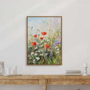 May include: A framed painting of a wildflower meadow, featuring red poppies, white daisies, and purple flowers. The artwork is set against a light blue sky, with a natural wood frame. The painting evokes a sense of a sunny meadow.