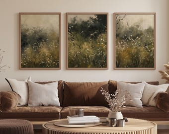 May include: Three framed landscape art prints. Each print features a field of wildflowers in shades of green, yellow, and white, with a soft, blurred effect. The frames are a light brown color. The artwork is displayed above a brown sofa with neutral-colored pillows.