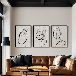 Puede incluir: Tres impresiones de arte abstracto en blanco y negro con líneas en marcos negros sobre una pared blanca encima de un sofá seccional de cuero marrón.