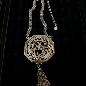 May include: A silver and gold tone necklace with a large octagonal pendant featuring a filigree design and a tassel. The pendant has a floral motif and a small bird in the center.