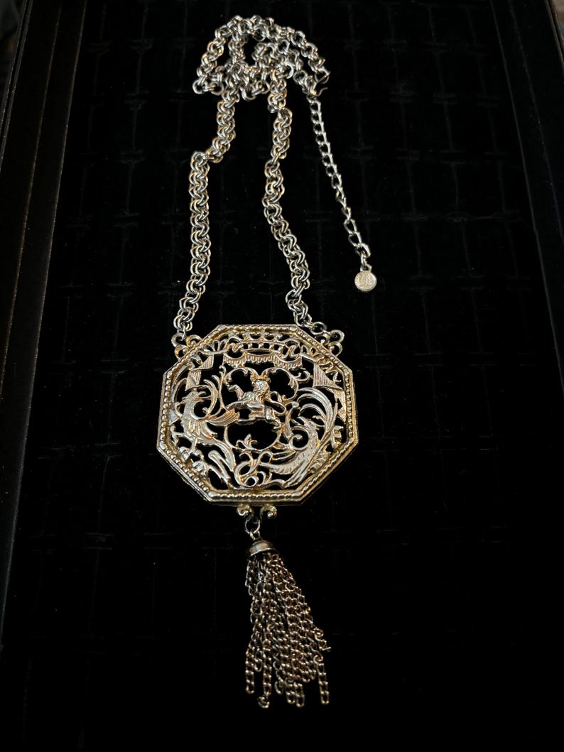 May include: A silver and gold tone necklace with a large octagonal pendant featuring a filigree design and a tassel. The pendant has a floral motif and a small bird in the center.