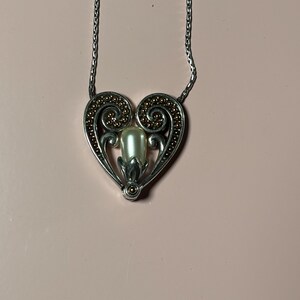 May include: Silver-toned necklace with a heart-shaped pendant. The pendant features a central pearl-like bead, surrounded by a decorative silver frame with gold-colored accents. The necklace chain is also silver.