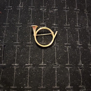 Puede incluir: Un broche de metal dorado en forma de cuerno de caza. El cuerno está enroscado en un círculo y tiene una campana en el extremo.