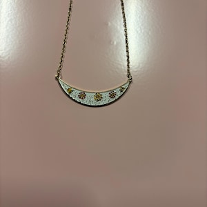 May include: A gold-toned necklace with a crescent-shaped pendant. The pendant is white with a floral design in yellow and red. The necklace hangs on a delicate gold chain, set against a pink background.