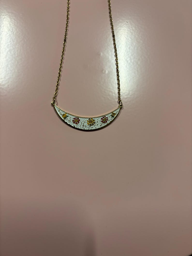 May include: A gold-toned necklace with a crescent-shaped pendant. The pendant is white with a floral design in yellow and red. The necklace hangs on a delicate gold chain, set against a pink background.