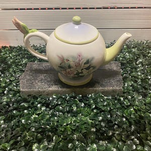 May include: A white ceramic teapot with a hummingbird design. The teapot has a green handle and a green hummingbird on the spout. The teapot is decorated with pink and white flowers.