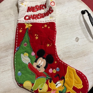May include: Red Christmas stocking with Mickey Mouse and Pluto. The stocking has a white cuff with "MERRY CHRISTMAS" embroidered in red. Mickey is decorating a Christmas tree. Pluto is lying down. The stocking is decorated with gold stars.