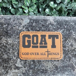 Pode incluir: Um patch retangular de couro castanho claro com a palavra "GOAT" em letras pretas em negrito. Abaixo, o texto "GOD OVER ALL THINGS" é exibido, junto com uma pequena cruz. O patch está sobre uma superfície cinza salpicada.