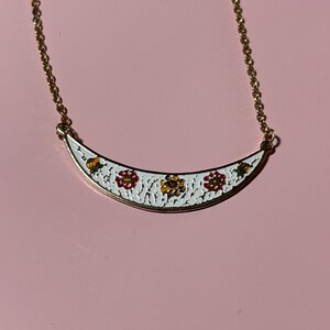 May include: A gold-toned necklace with a white, crescent-shaped pendant. The pendant features a floral design with red, yellow, and green accents. The necklace is displayed on a pink surface.