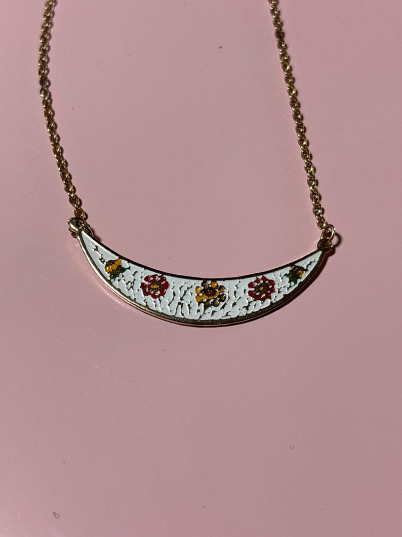 May include: A gold-toned necklace with a white, crescent-shaped pendant. The pendant features a floral design with red, yellow, and green accents. The necklace is displayed on a pink surface.