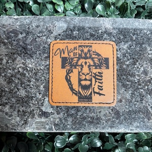 Könnte beinhalten: Ein quadratischer, hellbrauner Lederaufnäher mit den Worten "Man of Faith" und einem Kreuzdesign mit einem Löwengesicht. Der Aufnäher hat einen genähten Rand und ist vor einem dunklen, strukturierten Hintergrund platziert.