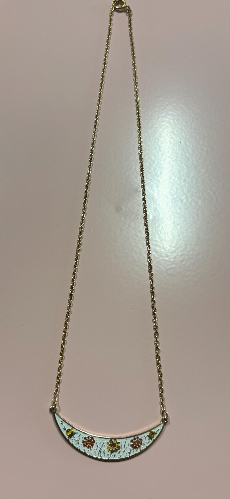 May include: Gold-toned necklace with a delicate chain and a curved pendant. The pendant features a white enamel base with a floral design in red and yellow. A simple and elegant piece of jewelry.