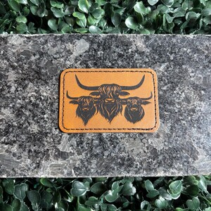 May include: A rectangular, tan leather patch featuring a black illustration of three Highland cattle. The patch has a stitched border and is set against a dark gray surface with green foliage.