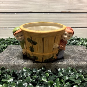 May include: A ceramic planter shaped like a barrel with two small figures peeking out from either side. The figures are painted with brown hair, green hats, and red and blue clothing. The planter is decorated with brown and green leaves.