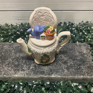 以下が含まれることがあります： 花柄の白い陶器製のティーポット。ティーポットの上には、小さな椅子があり、ジョウロ、植木鉢、小さな箱が置かれています。