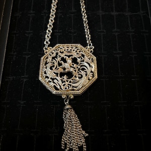 May include: A silver necklace with an octagonal pendant featuring an intricate filigree design and a tassel. The pendant has a floral motif and a small figure in the center.