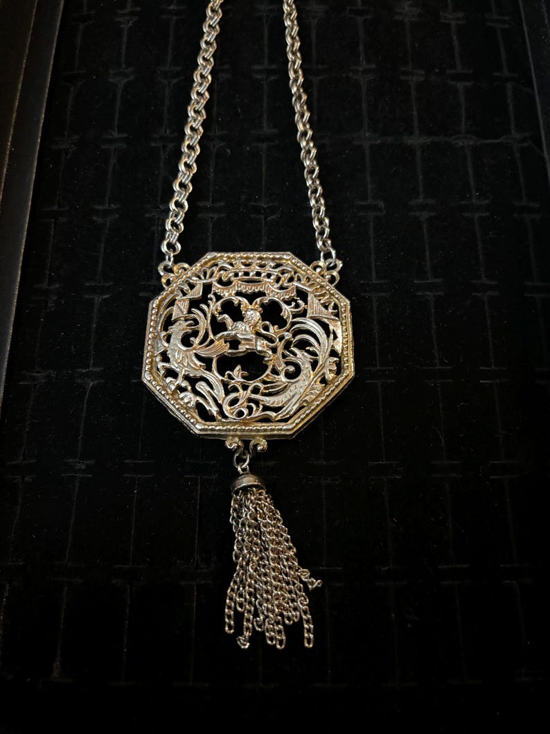 May include: A silver necklace with an octagonal pendant featuring an intricate filigree design and a tassel. The pendant has a floral motif and a small figure in the center.