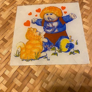 May include: A vintage glass Cabbage Patch Kids coaster featuring a cartoon image of a Cabbage Patch Kid in a blue and red outfit, with an orange cat, hearts, and flowers. The coaster is square and has a clear border.