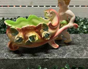 May include: A vintage ceramic planter featuring a cherub pushing a wheelbarrow. The wheelbarrow is painted in shades of pink, yellow, and green, with blue floral accents. The cherub has blonde hair and is nude. The planter is designed for small plants.