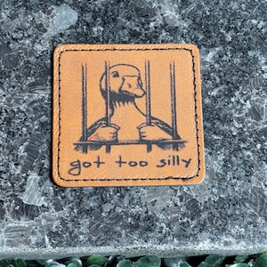 May include: A square, brown leather patch with a stitched border. The patch features a black illustration of a duck behind bars, with the words "got too silly" in a handwritten font.