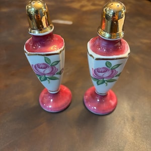 May include: A pair of vintage salt and pepper shakers. The shakers have a pink base and top with a white center section. Each shaker features a rose design and a gold-colored top.